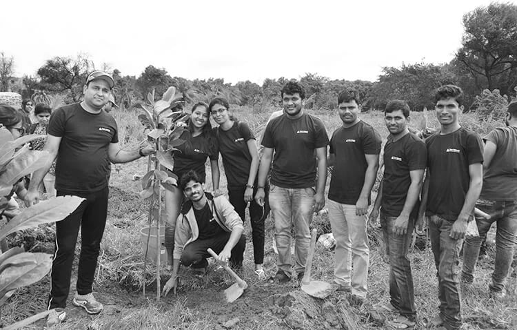 A circle of Tricon employees standing on a ground, clicking picture along with plants, depicting their engagement in social work