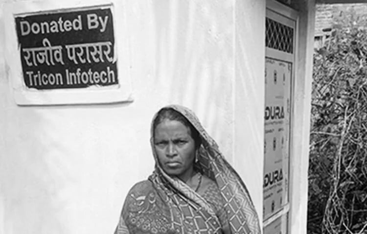 A woman centrally standing in the frame, with a board of donation by Tricon Infotech placed on the wall